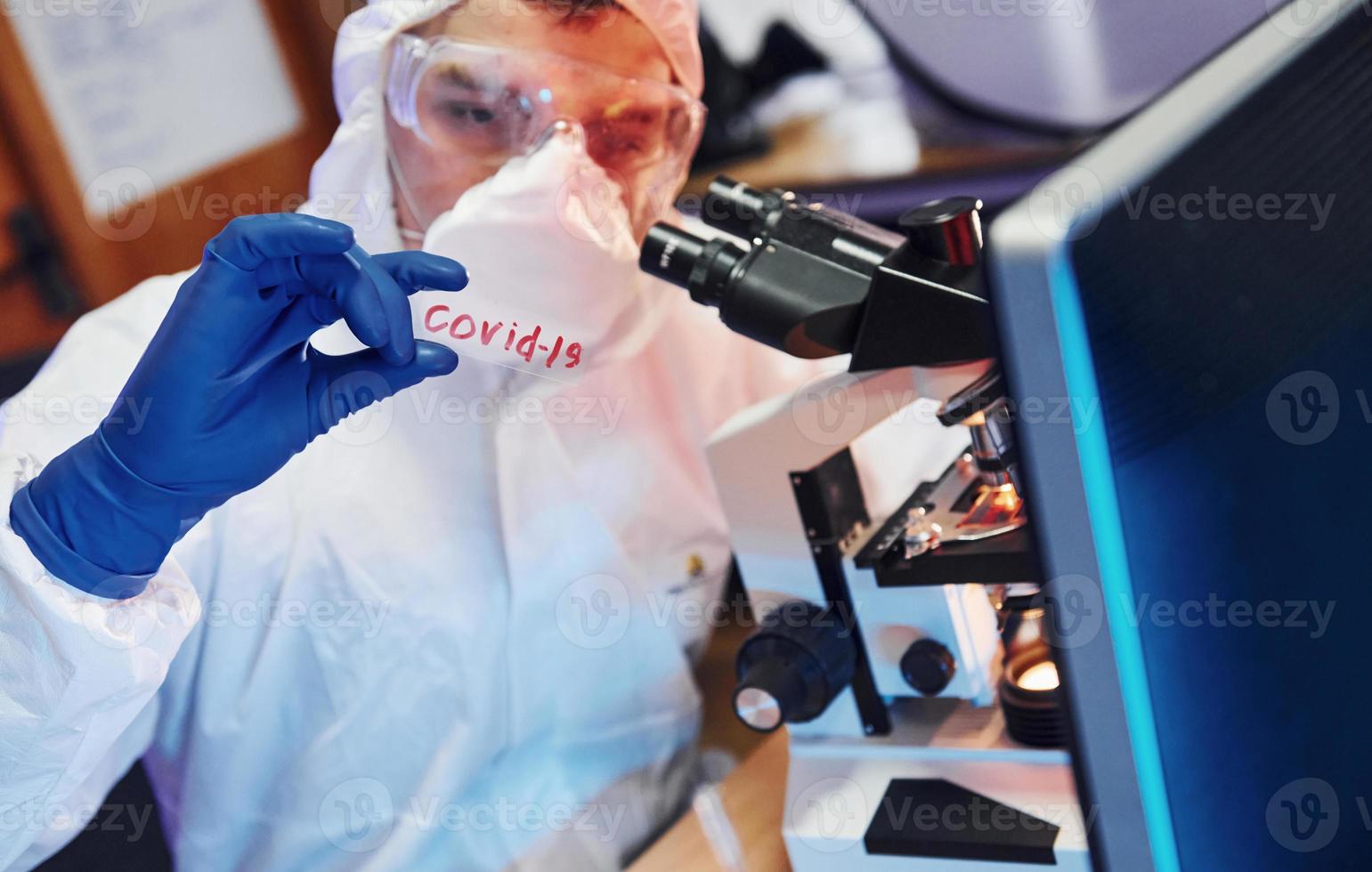 Scientist in white protective wear indoors in laboratory with lens with COVID-19 title on it photo