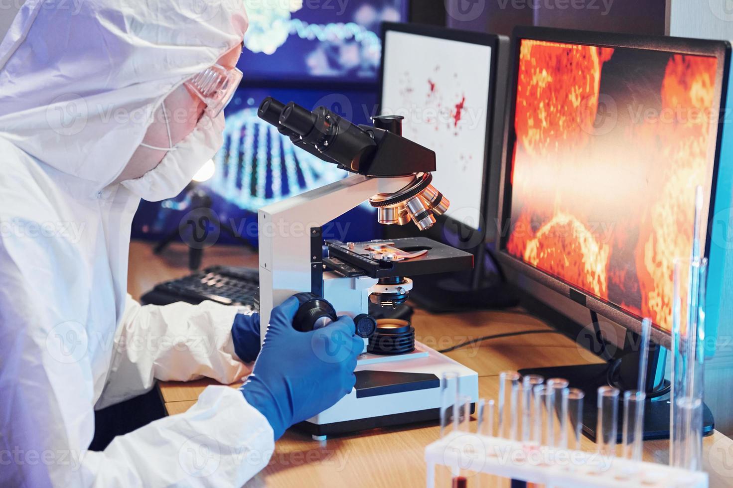 científico con uniforme protector blanco cerca de pantallas con datos trabaja con coronavirus y tubos de sangre en laboratorio foto