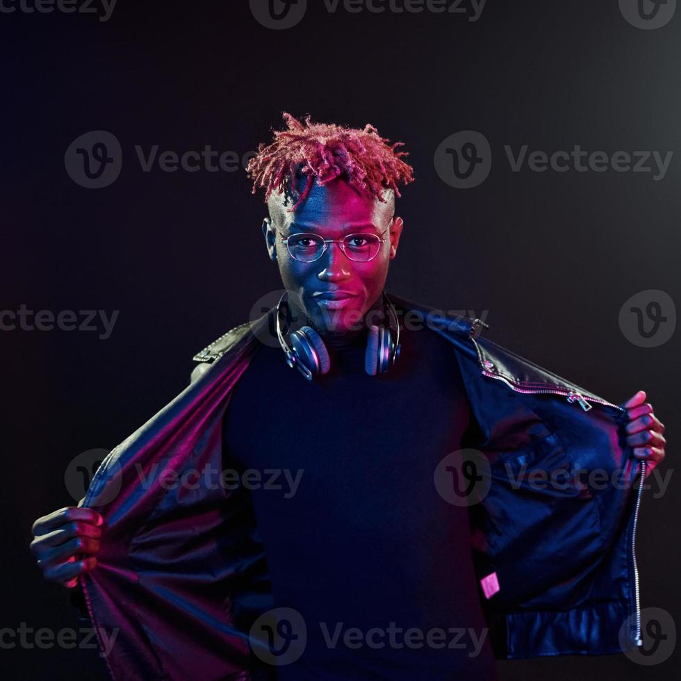 Posing with wireless headphones on neck. Futuristic neon lighting. Young african american man in the studio photo