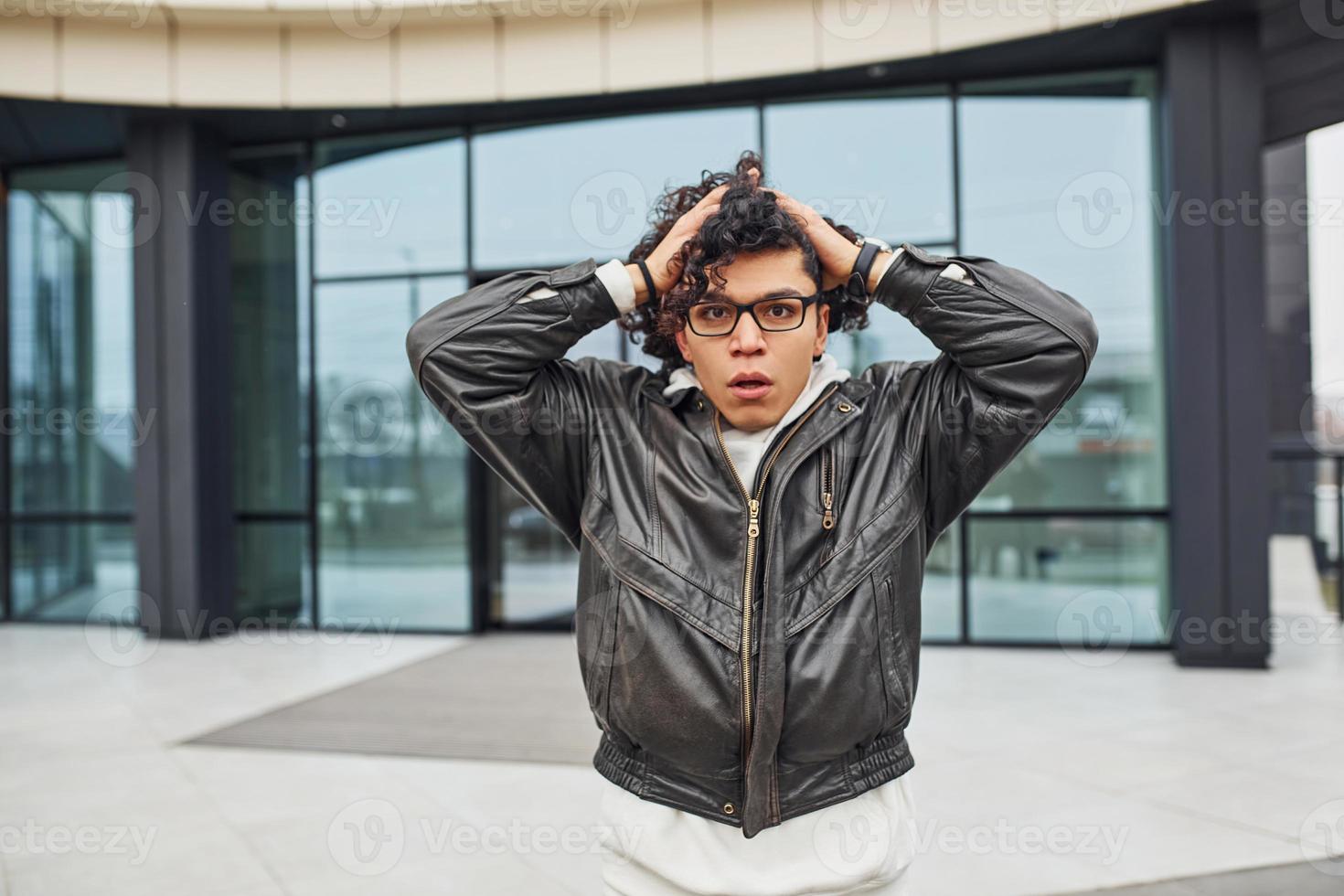 Handsome young man with curly black hair standing and feels shoked on the street against building photo