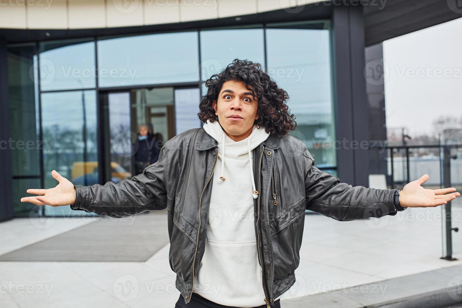 Handsome young man with curly black hair posing for the camera on the street against building photo