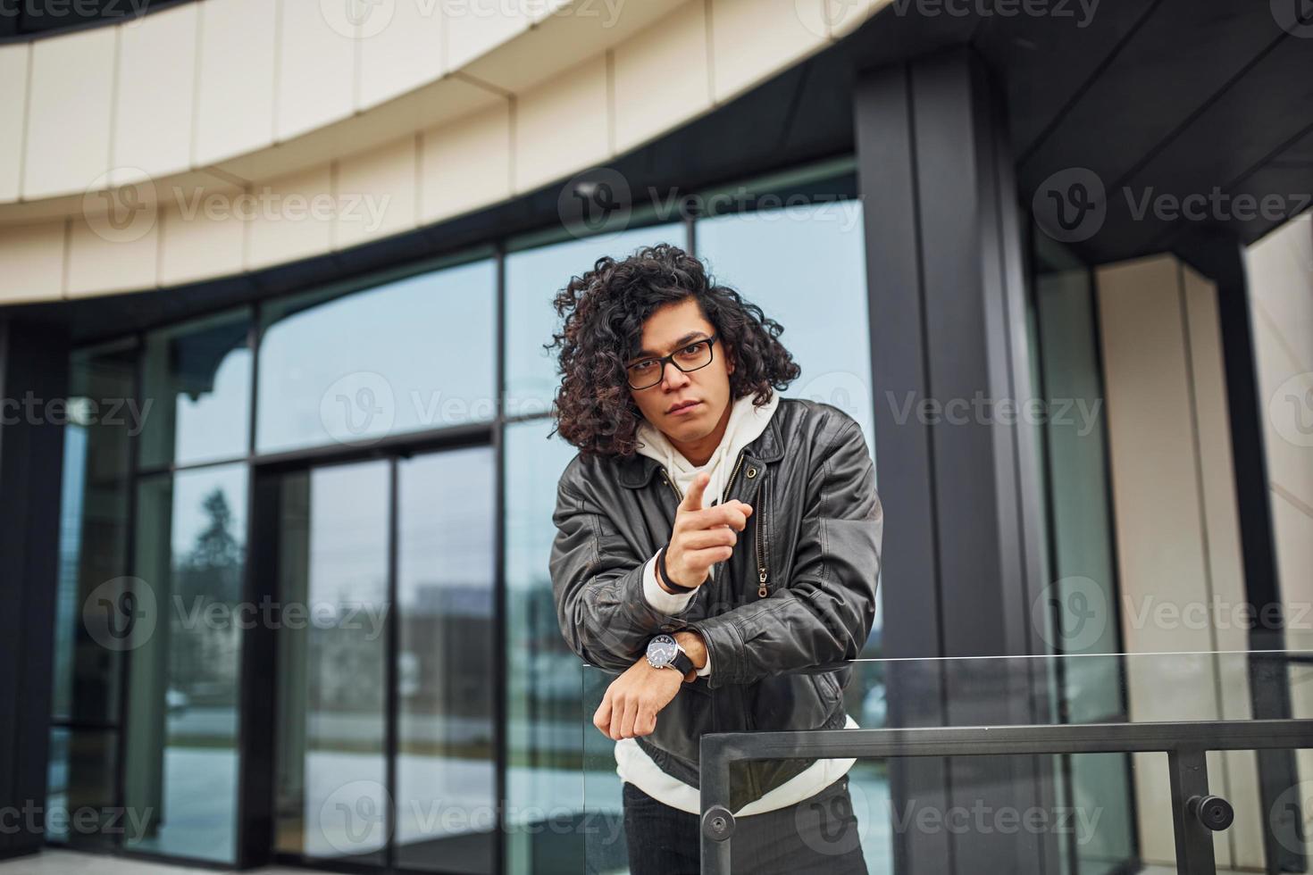 Handsome young man with curly black hair and in glasses is on the street against building photo