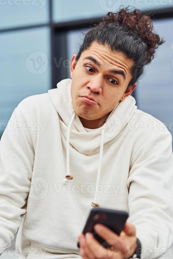 Handsome young man with curly black hair sitting on the street with smartphone against building photo