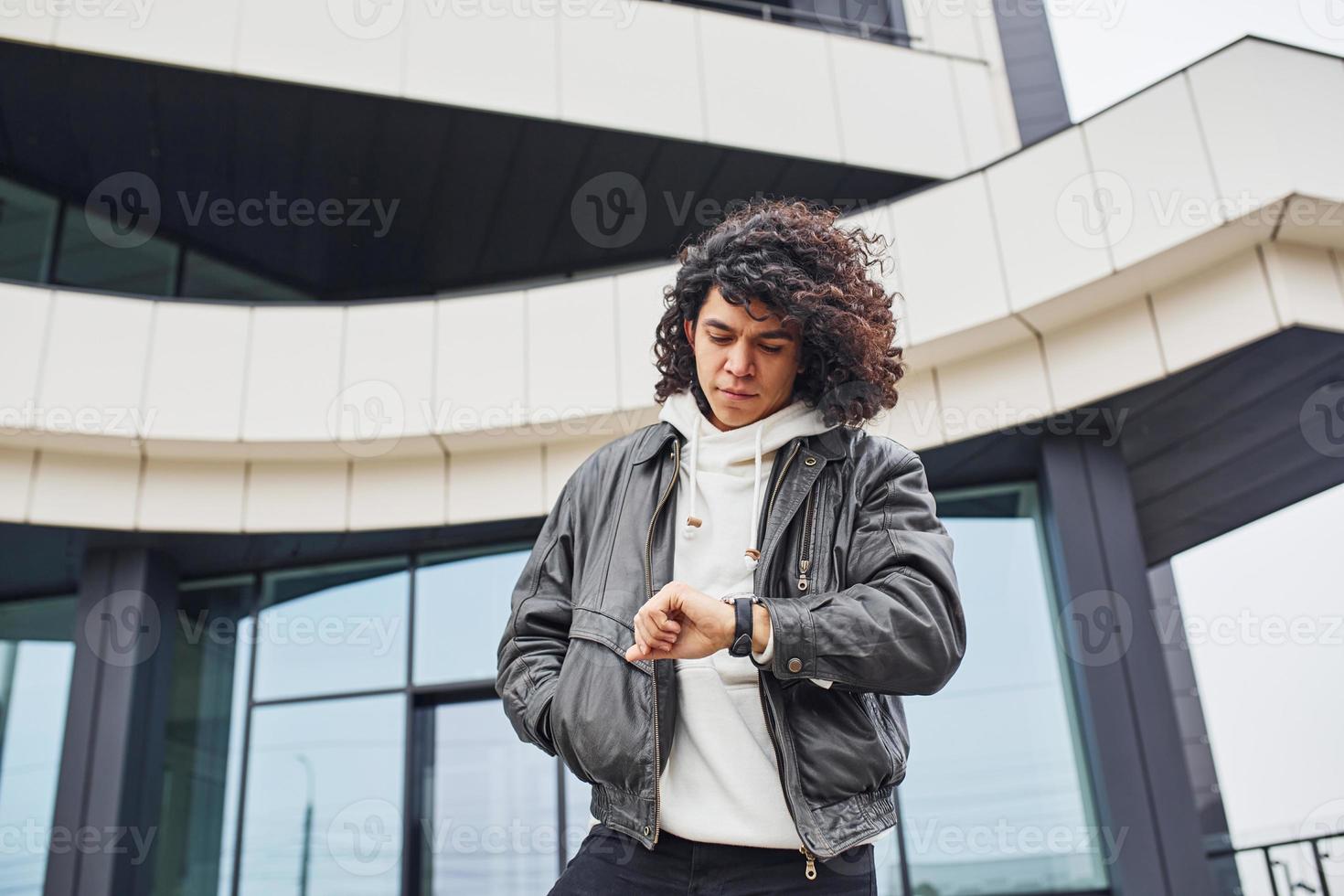 Handsome young man with curly black hair posing for the camera on the street against building photo
