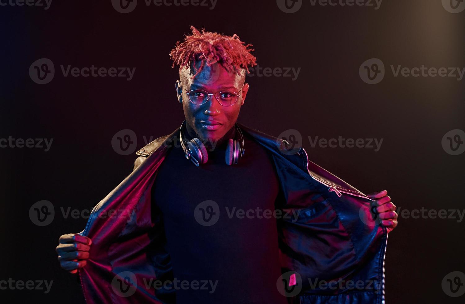 Posing with wireless headphones on neck. Futuristic neon lighting. Young african american man in the studio photo