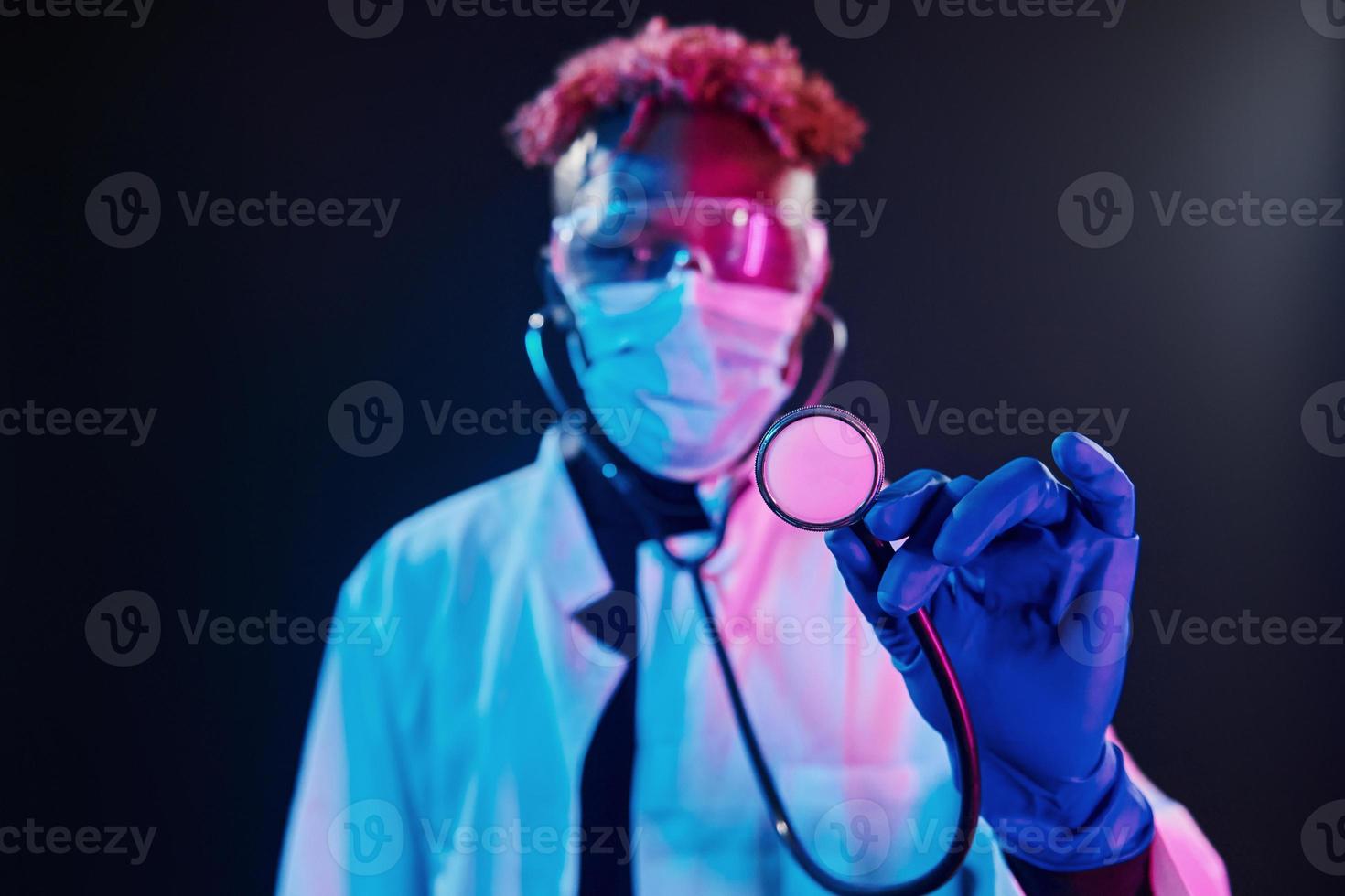 Smart doctor in protective uniform holding stethoscope. Futuristic neon lighting. Young african american man in the studio photo