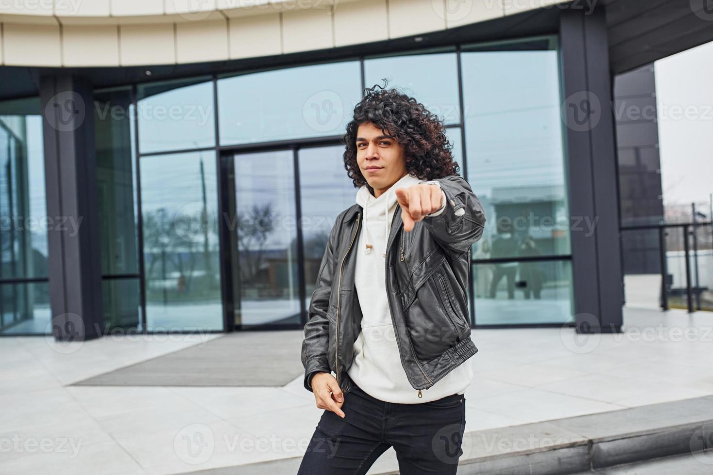 Handsome young man with curly black hair posing for the camera on the street against building photo