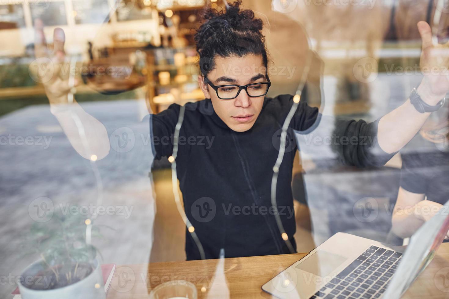 Young guy with black curly hair sitting indoors in cafe with laptop. View through the glass photo