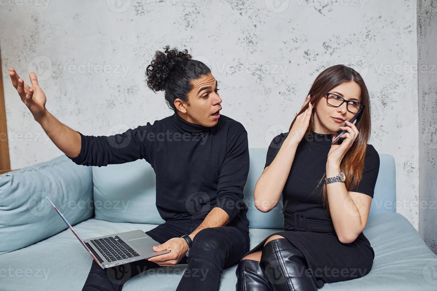 Man with curly hair and woman that in black clothes sitting with laptop indoors. Guy is angry photo