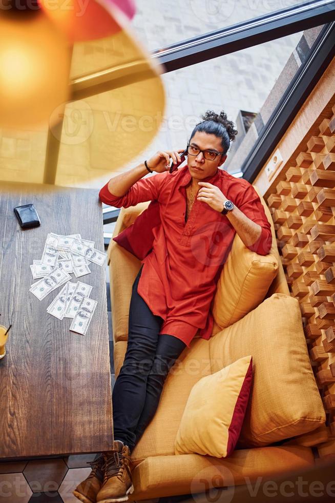 Rich man in red and black clothes sitting indoors in cafe with lot of money on the table photo