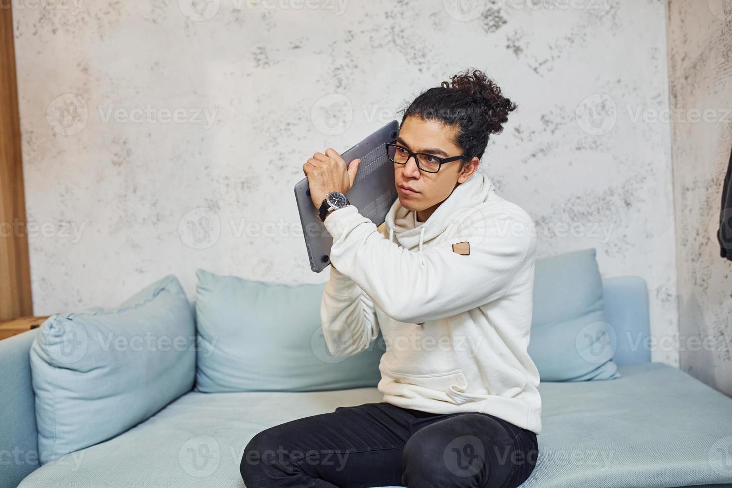 Angry young man with curly black hair goes crazy and angry with laptop in hands photo