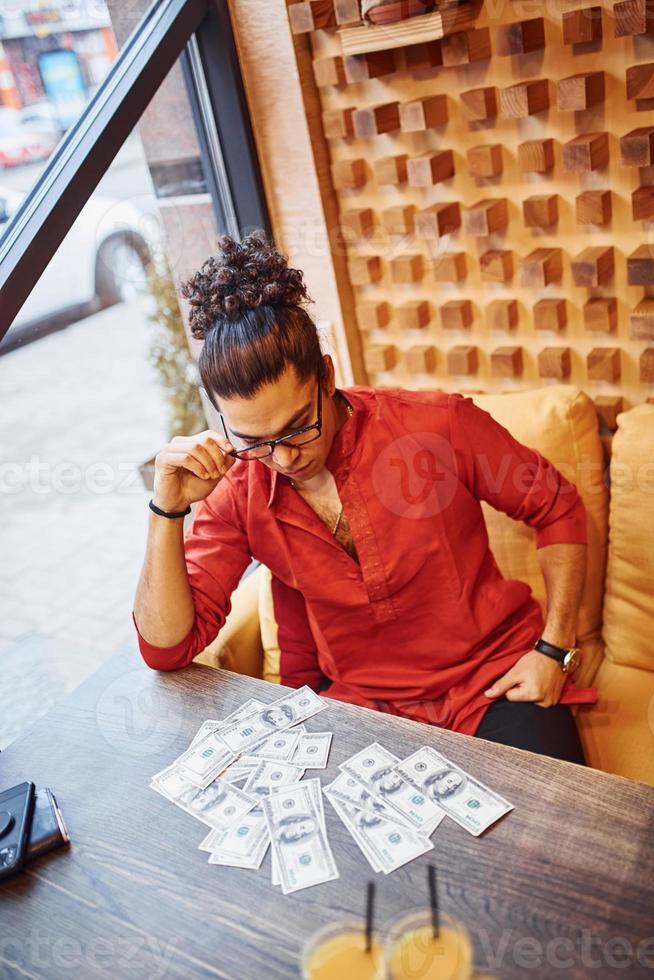 Rich man in red and black clothes sitting indoors in cafe with lot of money on the table photo