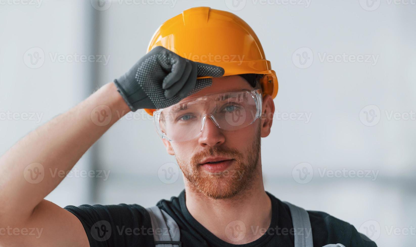 Portrait of man in grey uniform, protective glasses and orange hard hat that standing indoors in modern big office at daytime photo