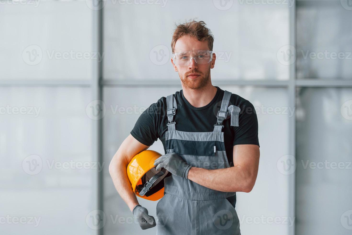 Portrait of man in grey uniform andwith orange hard hat in hands that standing indoors in modern big office at daytime photo