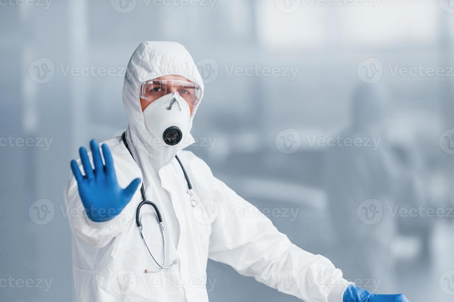 muestra el gesto de parada a mano. médico científico masculino en bata de laboratorio, anteojos defensivos y máscara foto