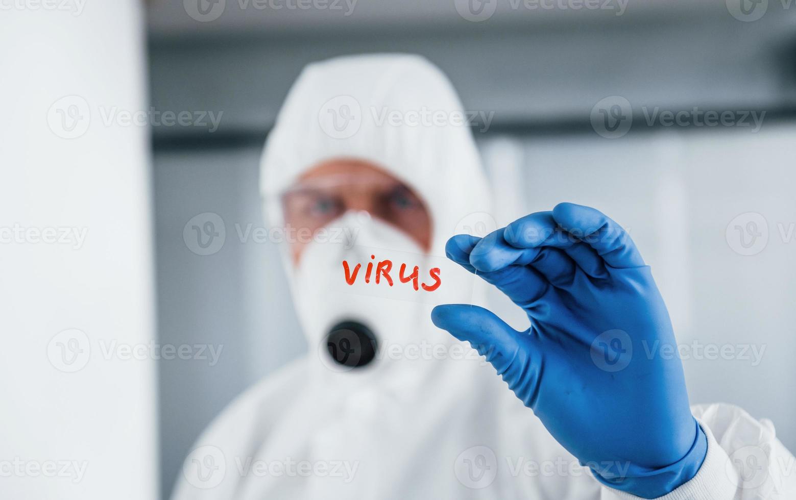 médico científico masculino en bata de laboratorio, anteojos defensivos y máscara sostiene vidrio con palabra de virus en él foto
