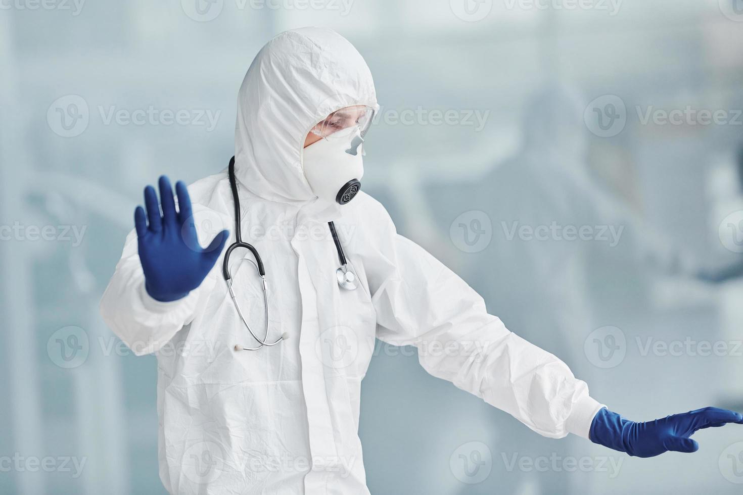 Shows stop gesture by hand. Male doctor scientist in lab coat, defensive eyewear and mask photo