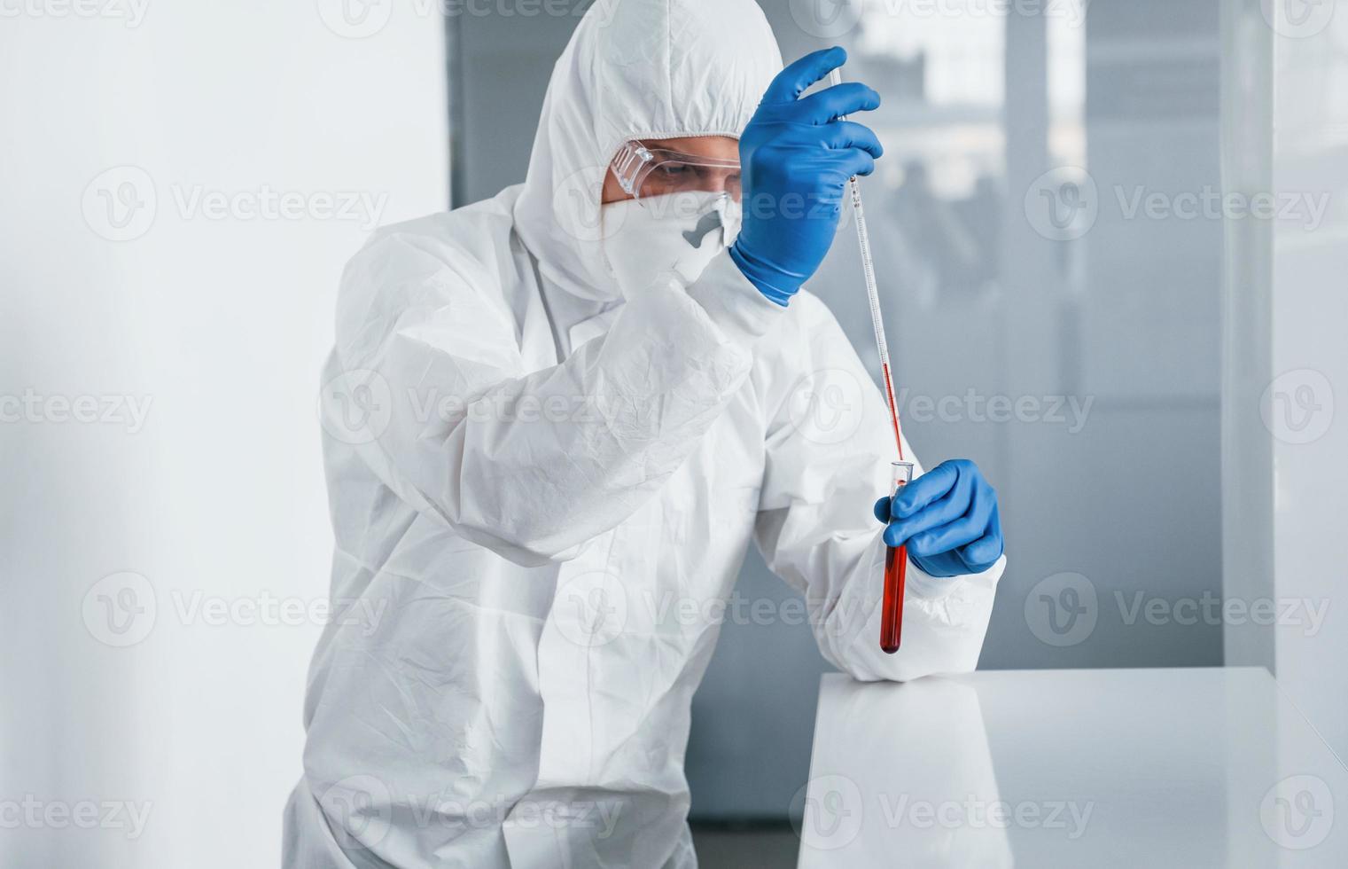 Male doctor scientist in lab coat, defensive eyewear and mask works with test tube with blood inside of it photo