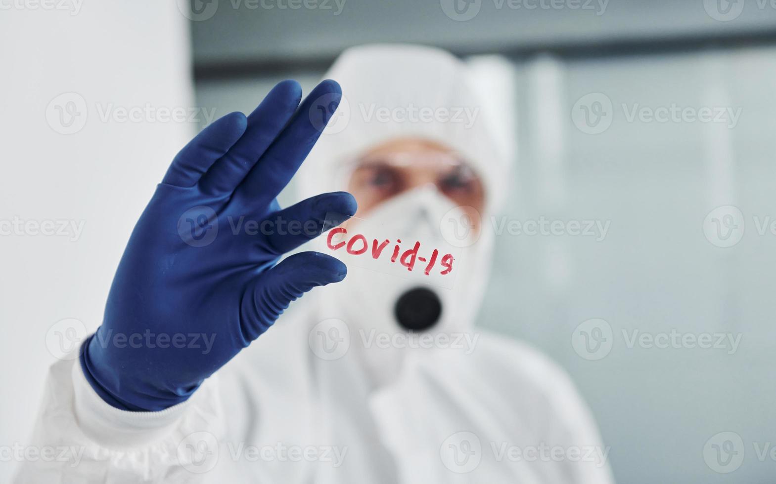 Male doctor scientist in lab coat, defensive eyewear and mask holds glass with covid-19 word on it photo