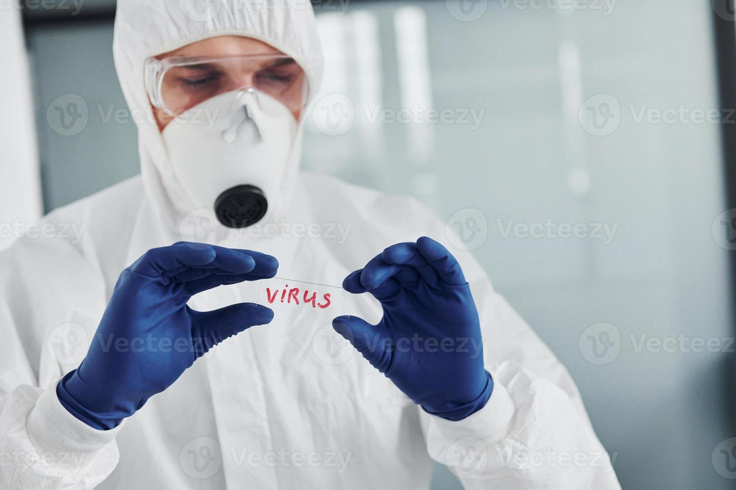 Male doctor scientist in lab coat, defensive eyewear and mask holds glass with virus word on it photo