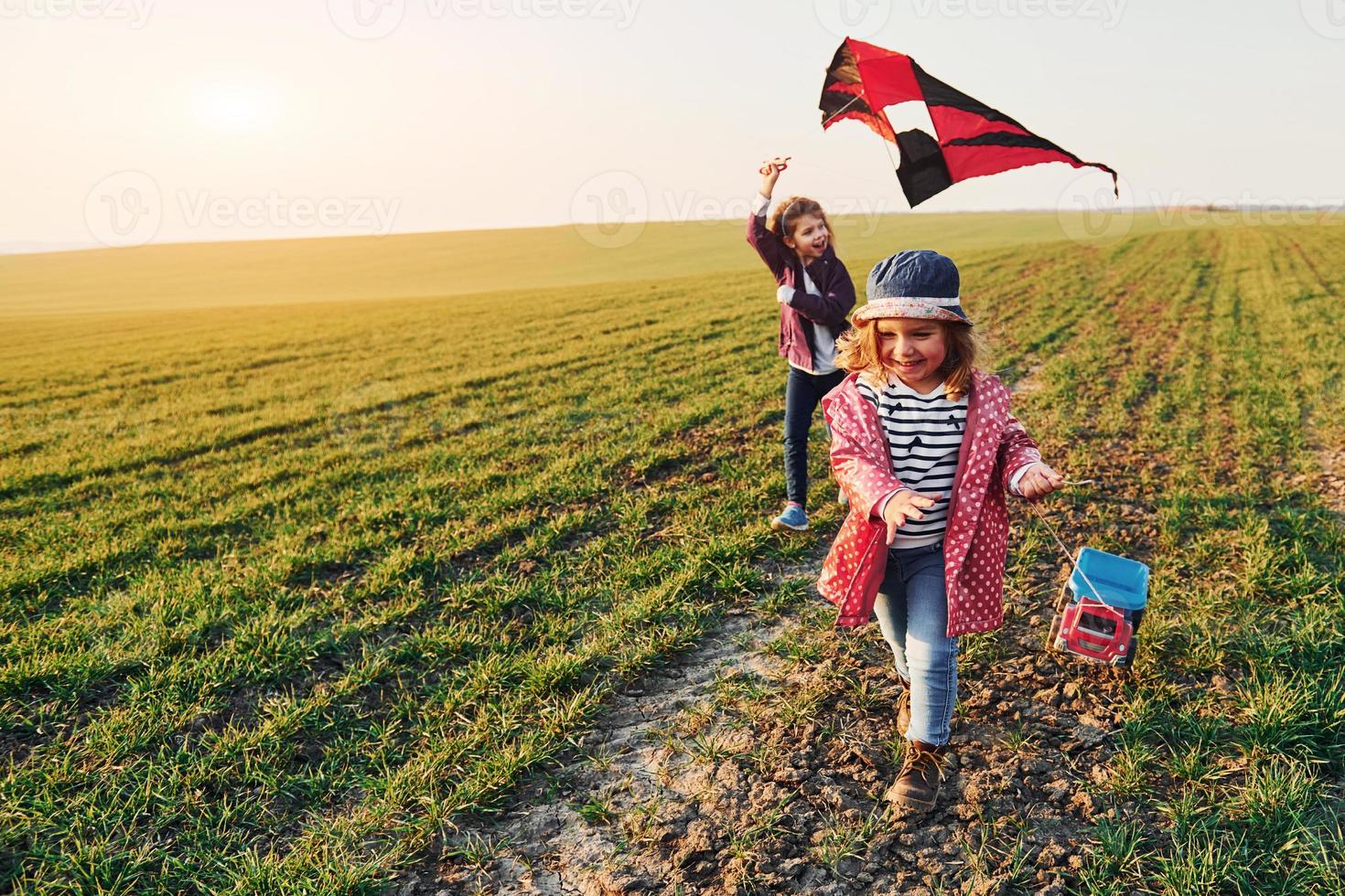 Two little girls friends have fun together with kite and toy car on the field at sunny daytime photo