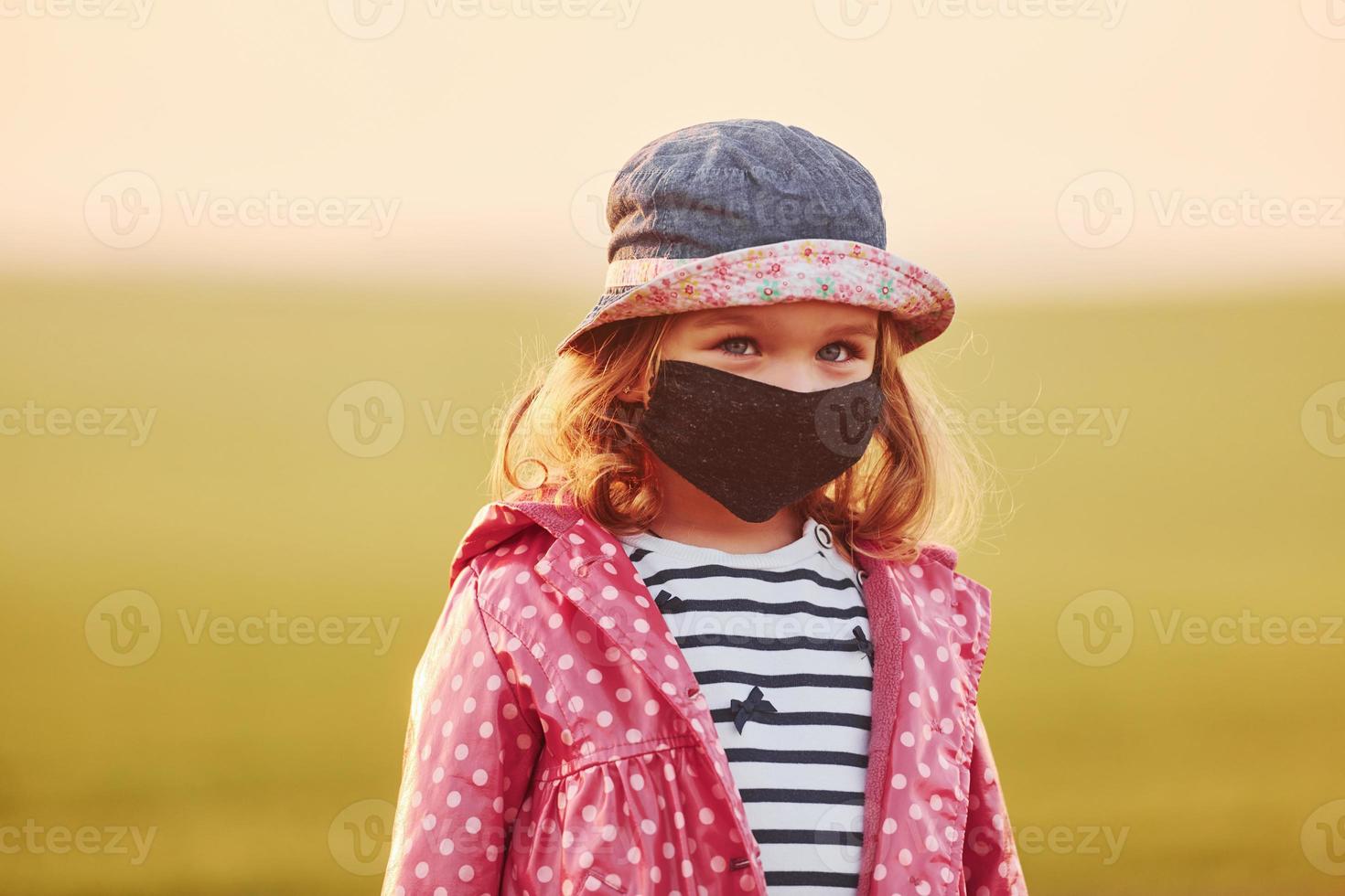 Portrait of cute little girl in black protective mask that standing outdoors illuminated by orange sunlight photo