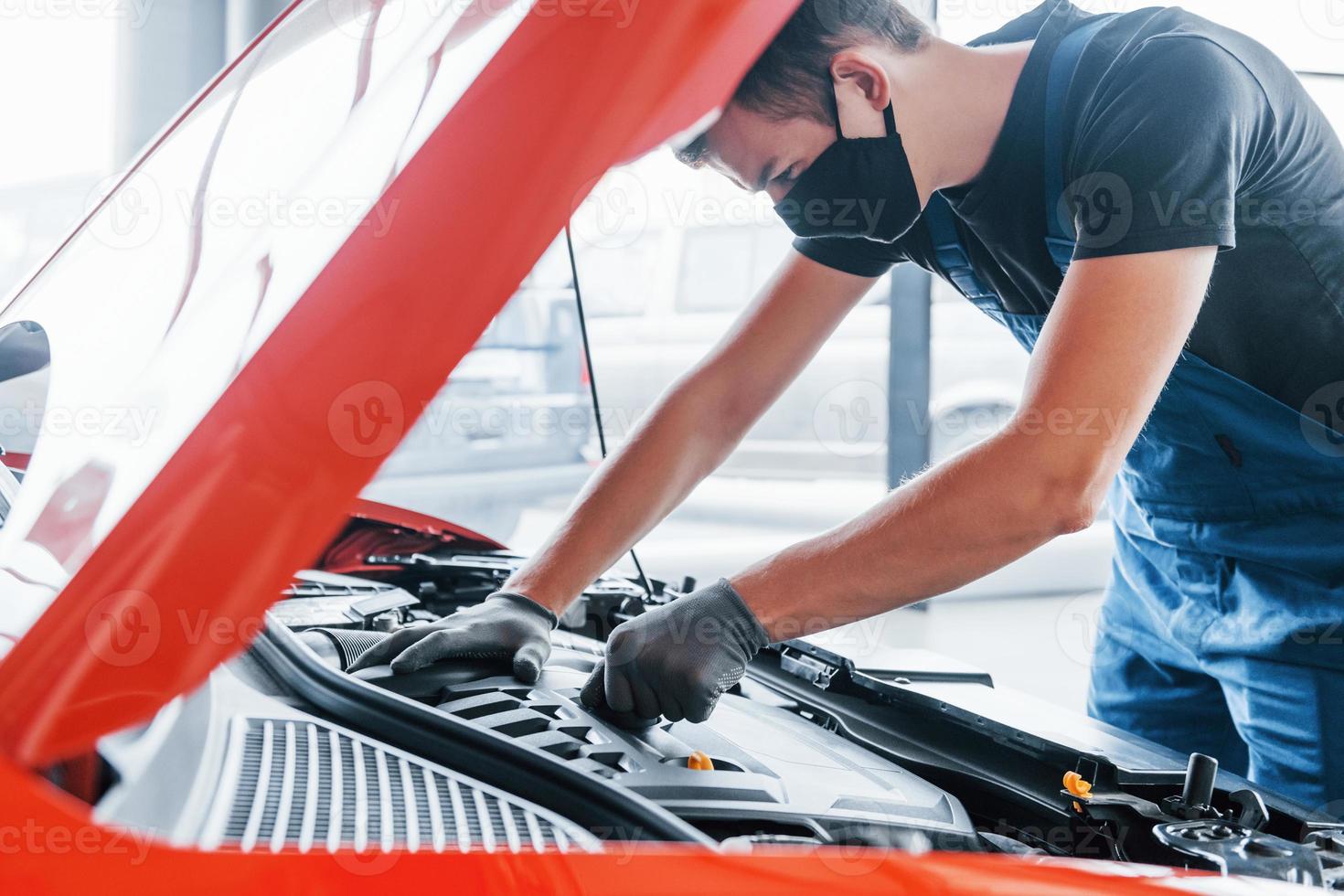 Man in uniform and black protective mask works with broken automobile. Conception of car service photo