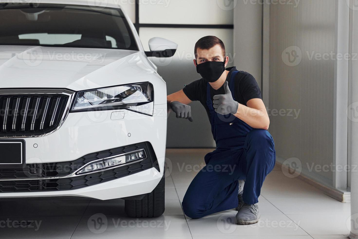 Guy in blue uniform and black protective mask sitting near fixed white car and shows thumb up photo