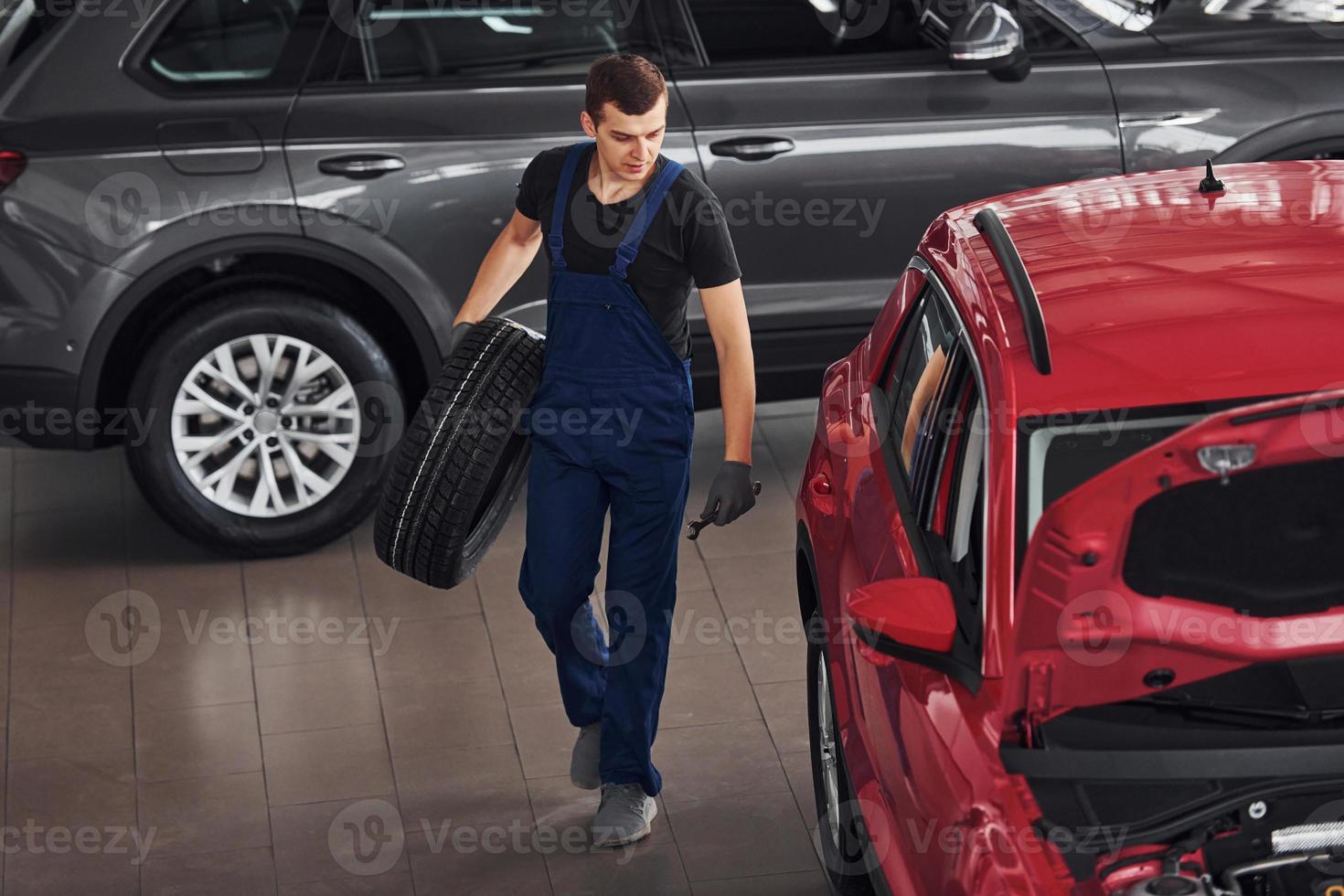 el trabajador con uniforme negro y azul sostiene la rueda del auto y camina con un automóvil cerca de rojo foto