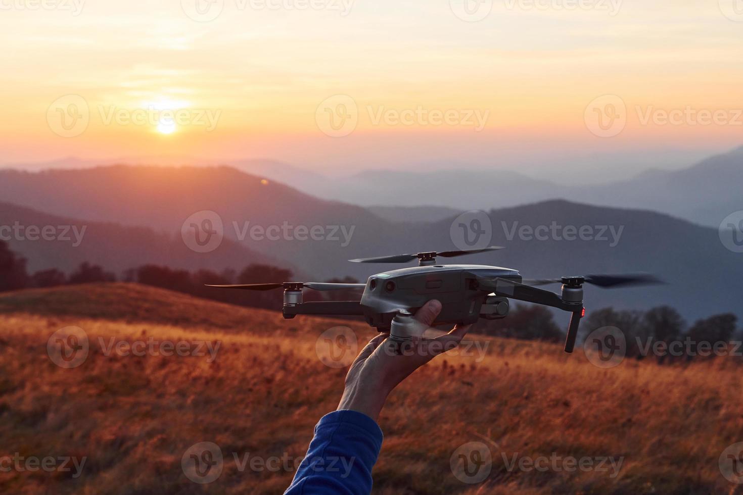 Man standing in the autumn field and holds drone in hand. Beautiful sunrise. Majestic landscape of mountains far away photo