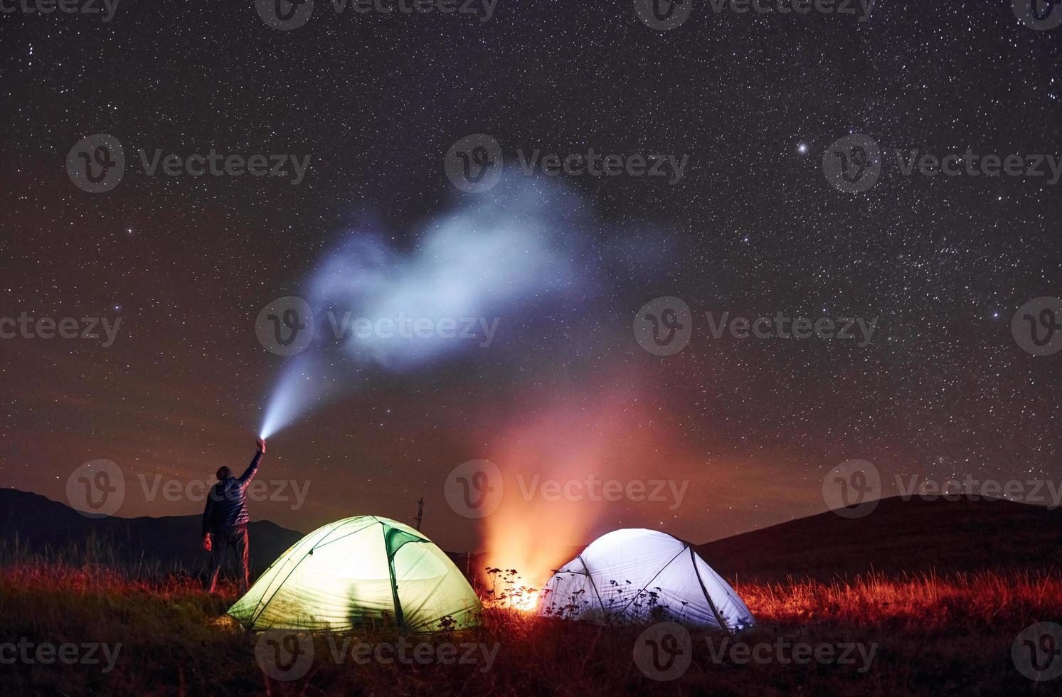 Man holds device with light beam. Two iluminated tents with campfire under stars at mountains at night photo