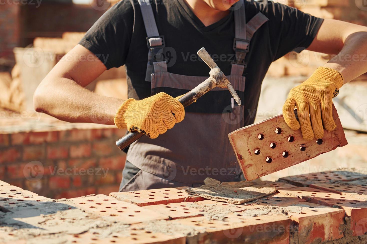 Holding brick and using hammer. Construction worker in uniform and safety equipment have job on building photo