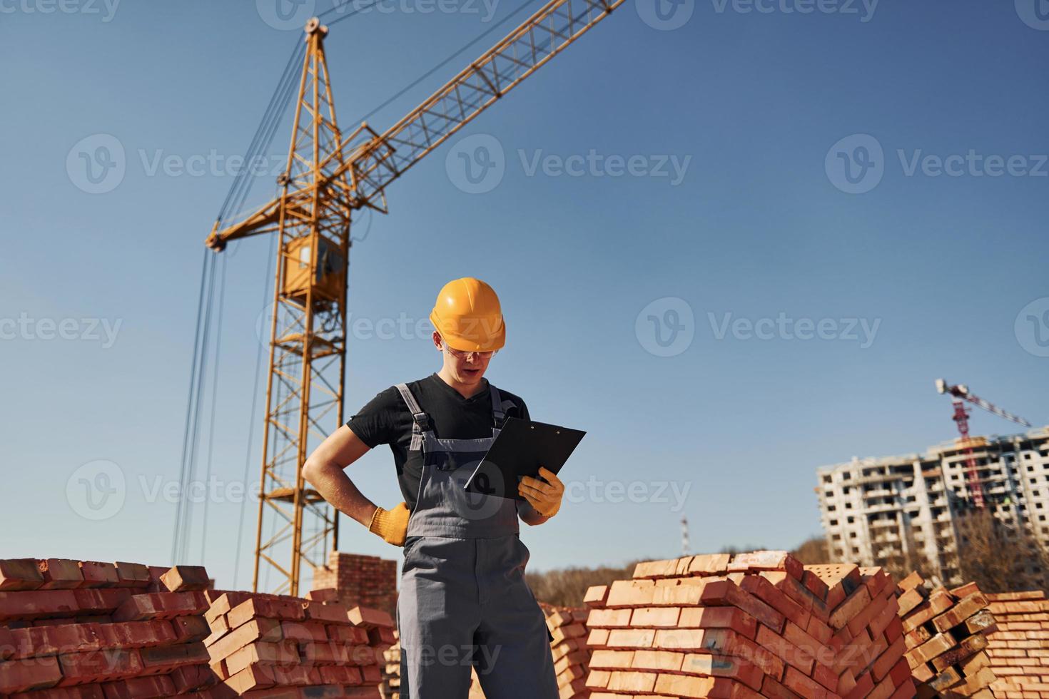 Holds notepad with documents. Construction worker in uniform and safety equipment have job on building photo