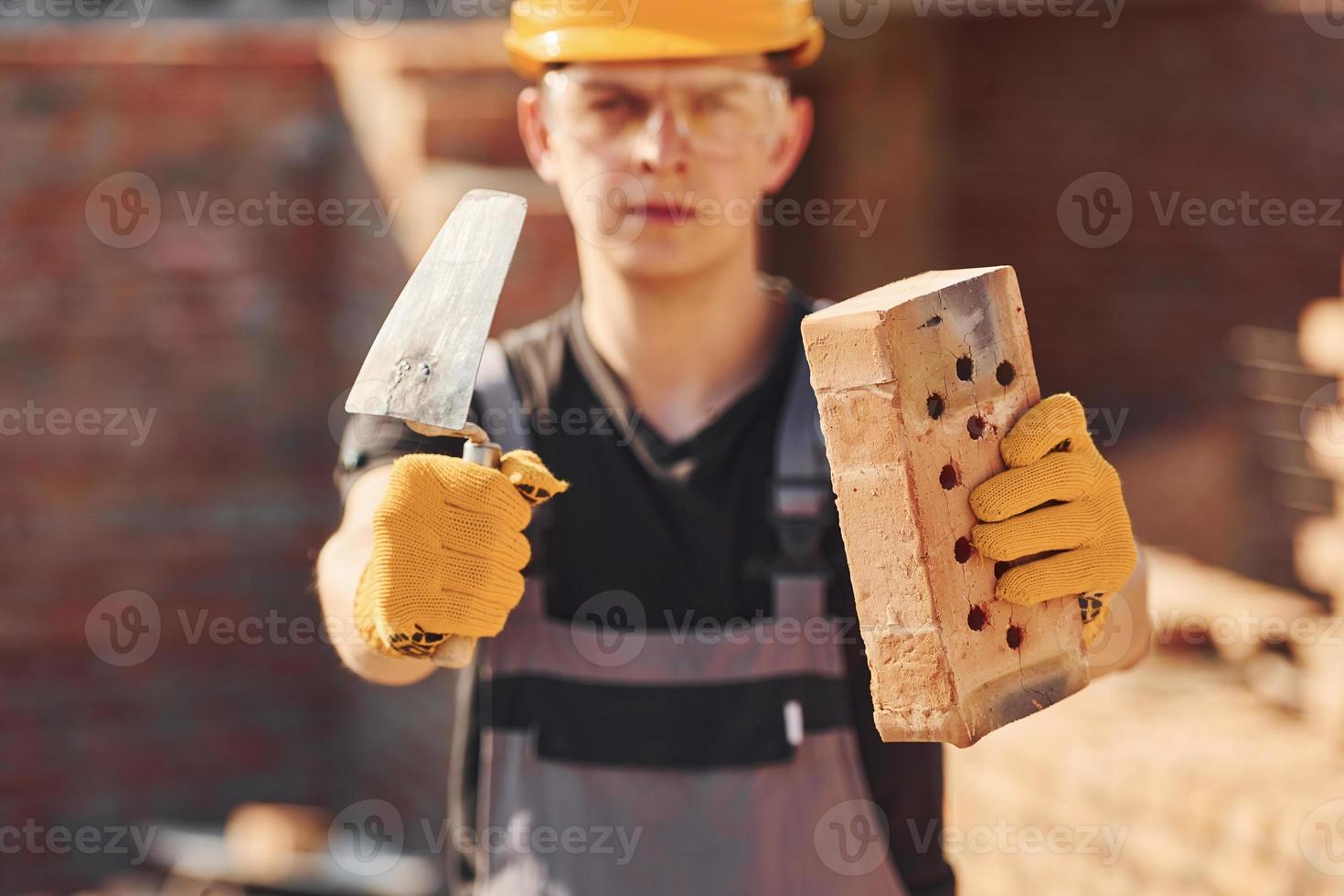 Portrait of construction worker in uniform and safety equipment that standing on building and holding brick and tool photo