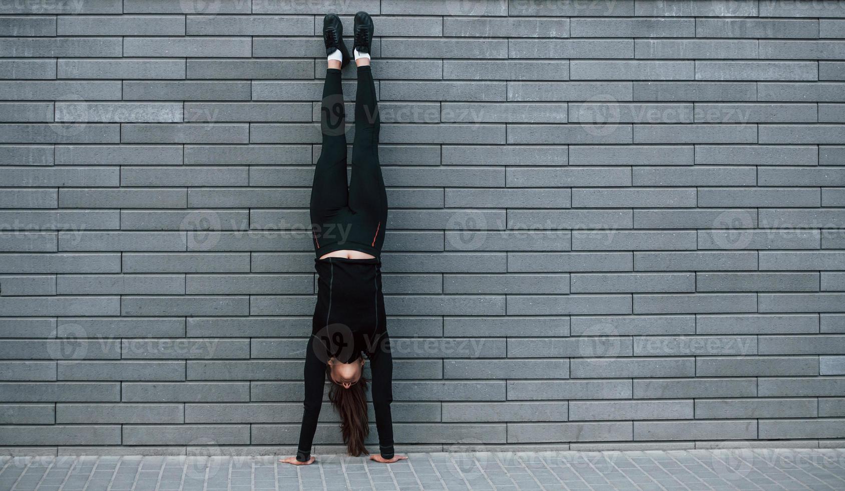 joven deportista con ropa deportiva negra haciendo ejercicios de parada de manos duros al aire libre cerca de la pared gris foto