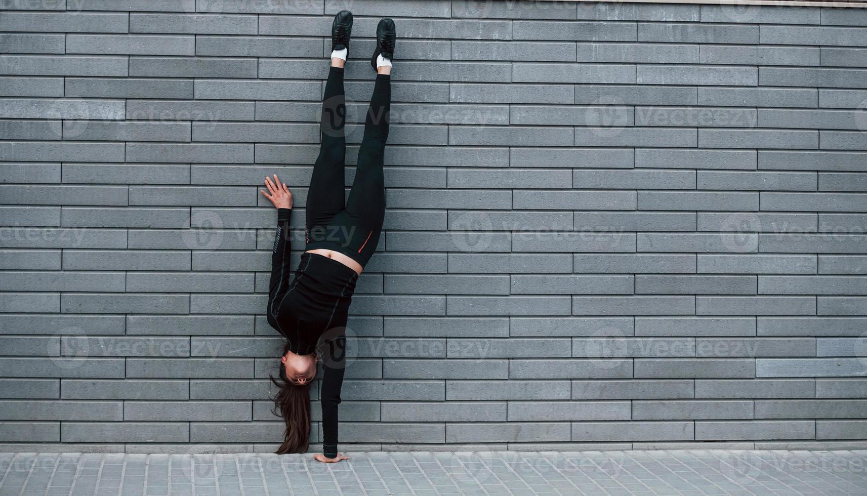 Young sportive girl in black sportswear doing hard handstand exercises outdoors near gray wall photo