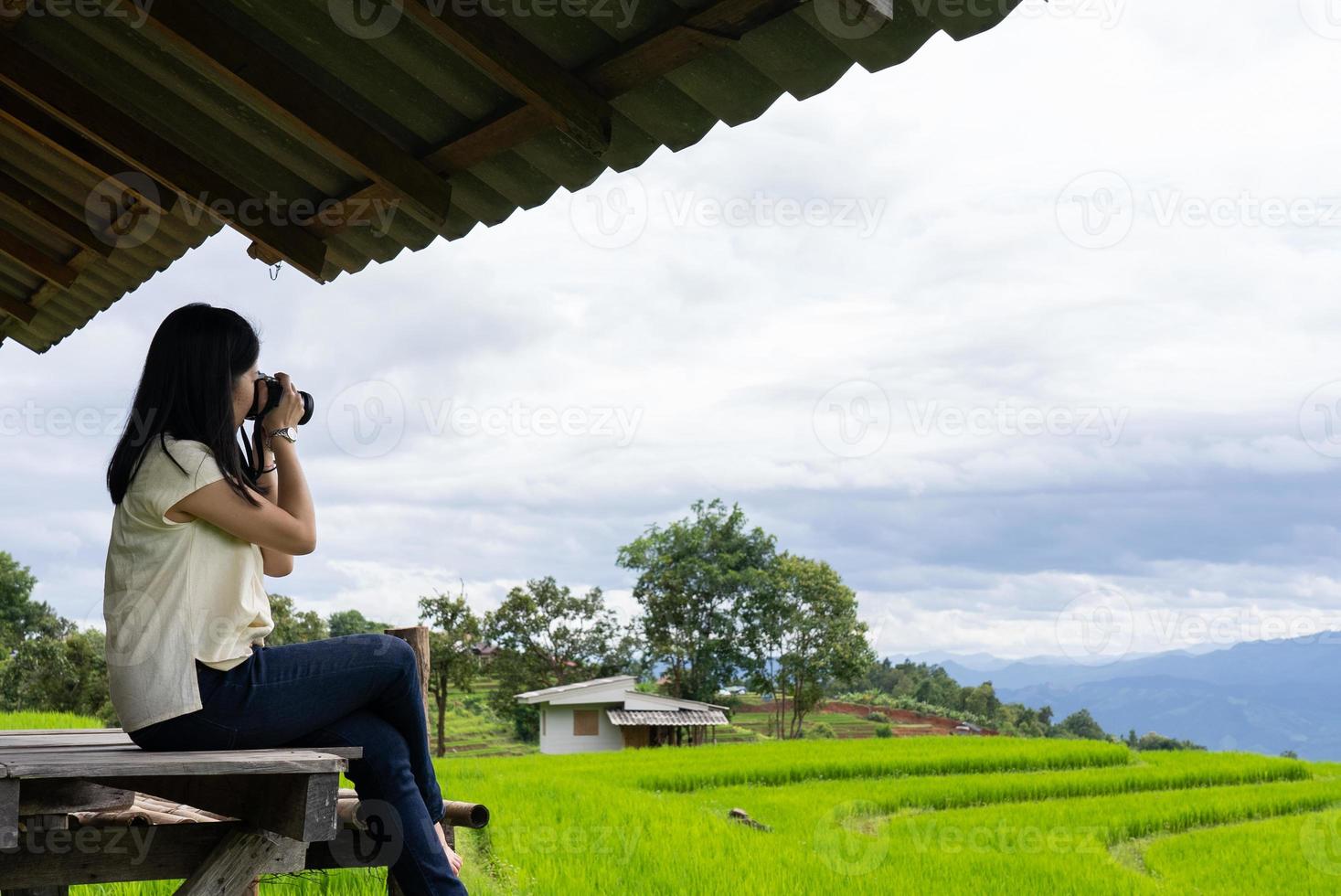 mujer sentada en la terraza de una pequeña cabaña en el pueblo alrededor del campo de arroz verde en temporada de lluvias. foto