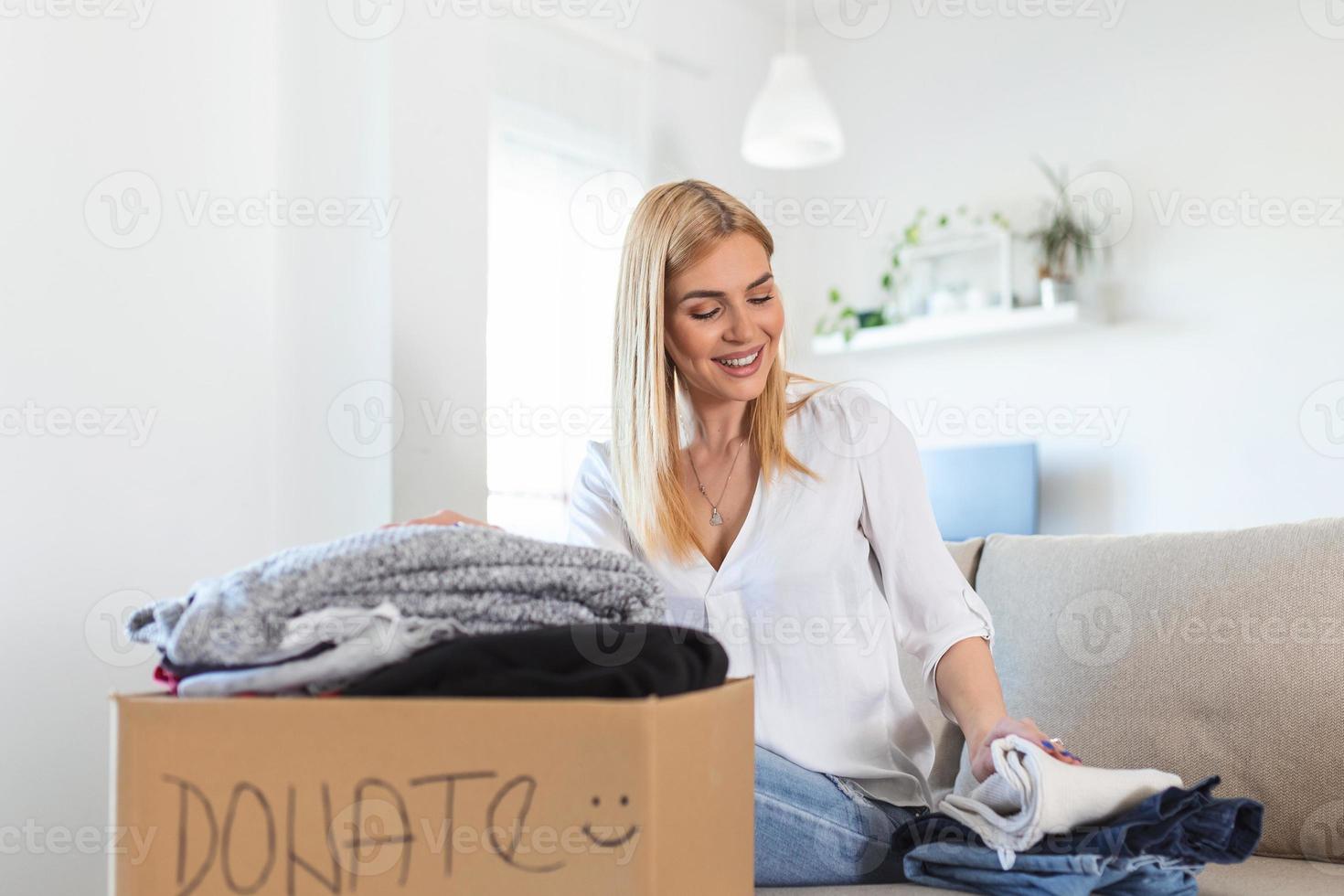 Happy young woman sit on couch stuck clothes in donation box at home, caring biracial female volunteer put apparel in carton package, donate to needy people, reuse, recycle concept photo