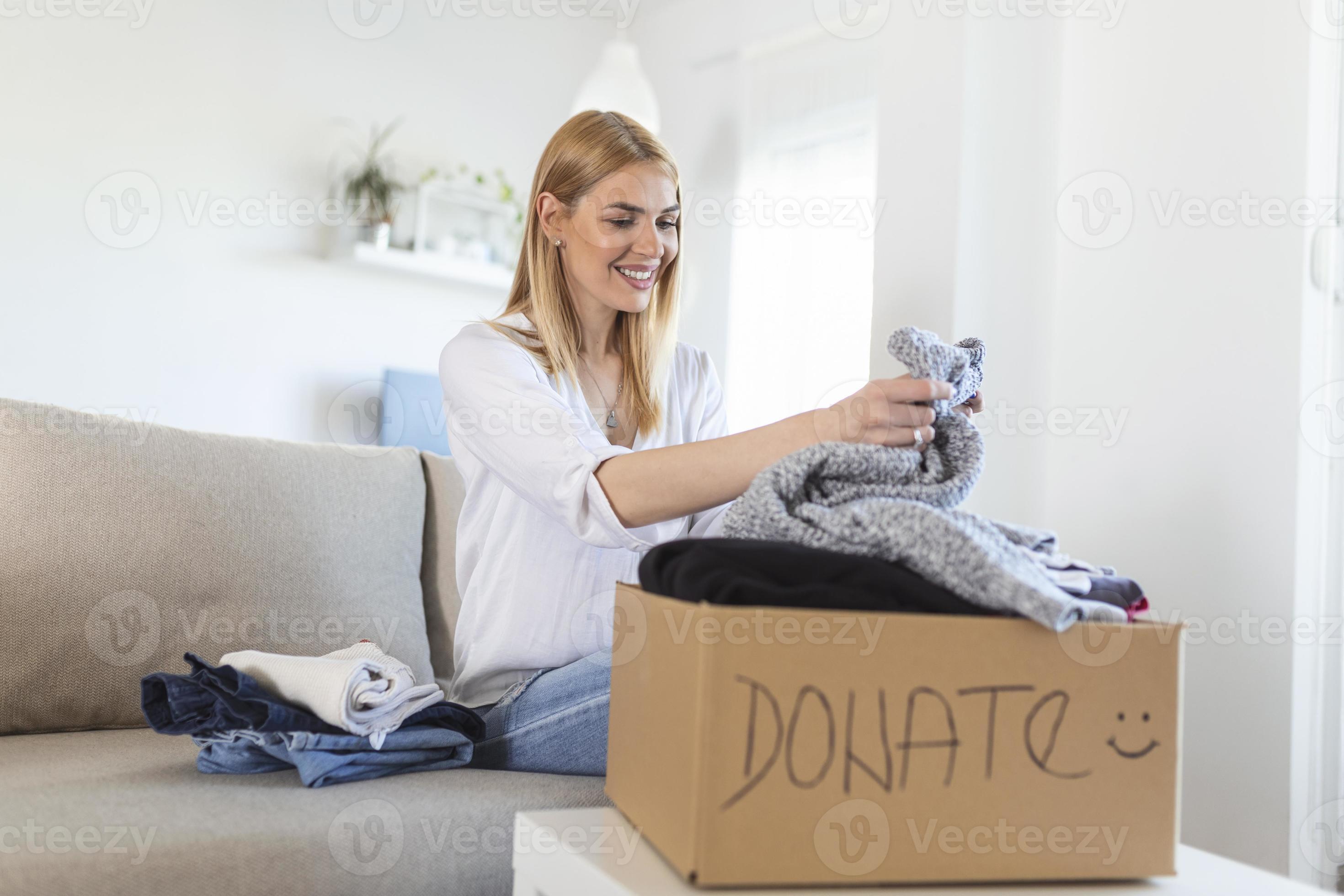Happy young woman sit on couch stuck clothes in donation box at home