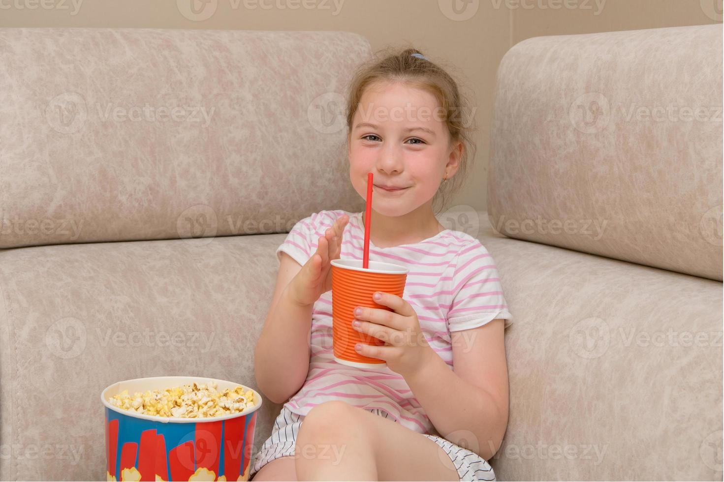 cute girl with a pigtail joyfully drinks cola and eats popcorn while sitting on the couch at home photo