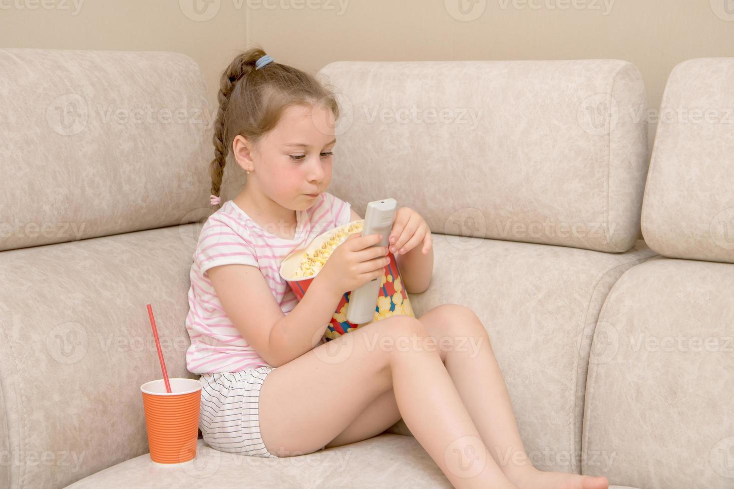 linda chica en casa en el sofá con un gran vaso de palomitas de maíz y coca cola tratando de descifrar el control remoto para cambiar los canales de televisión foto