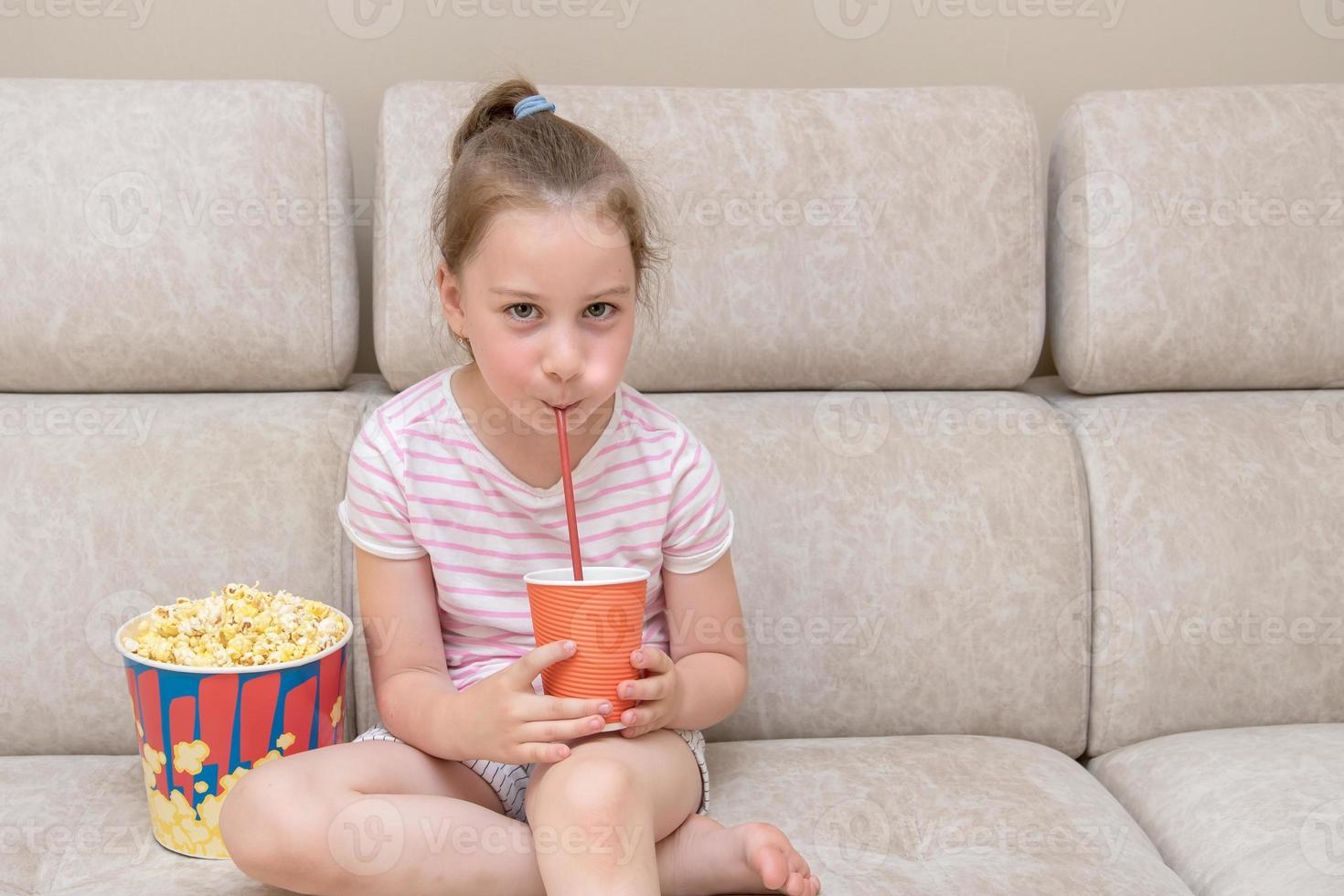 cute little girl sits on the couch at home and drinks a cocktail from a straw, next to it is a huge glass of popcorn photo