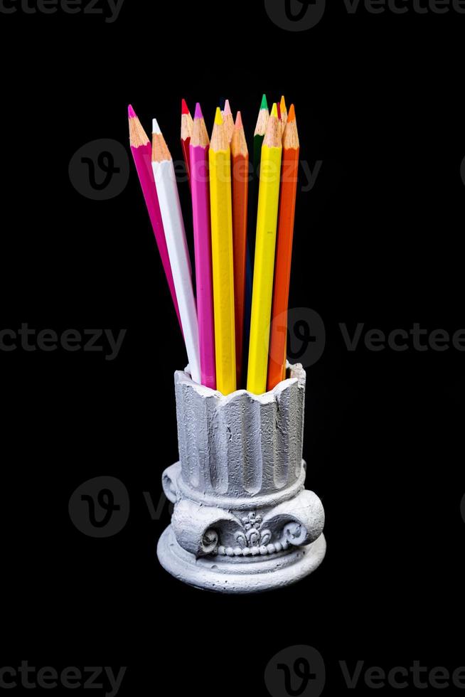 Color pencils for the drawing, located in a support as a vase, focus in front. multi-colored pencils and felt-tip pens in a plaster vase in the shape of a antique columnon a black background. photo