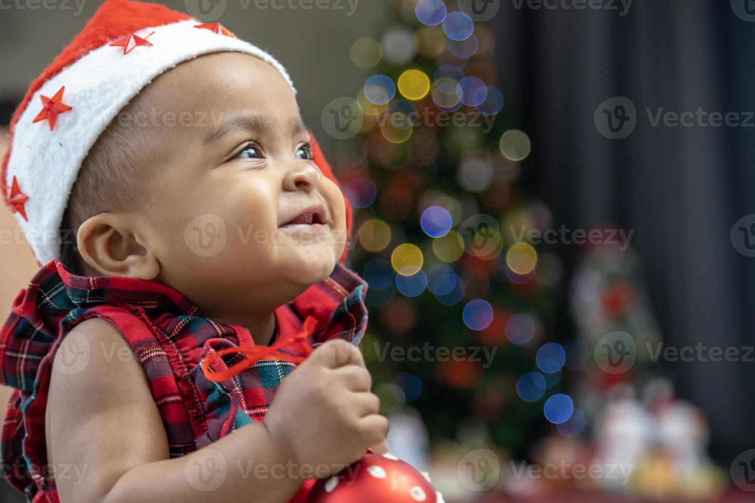 African American baby is happily smiling as playing with ornament bauble while dressing in christmas dress and santa hat with christmas tree on the back for season celebration photo