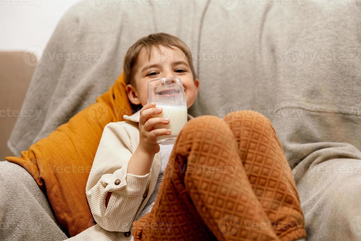 Little adorable boy sitting on the couch at home and drinking milk. Fresh milk in glass, dairy healthy drink. Healthcare, source of calcium, lactose. Preschool child with casual clothing. photo