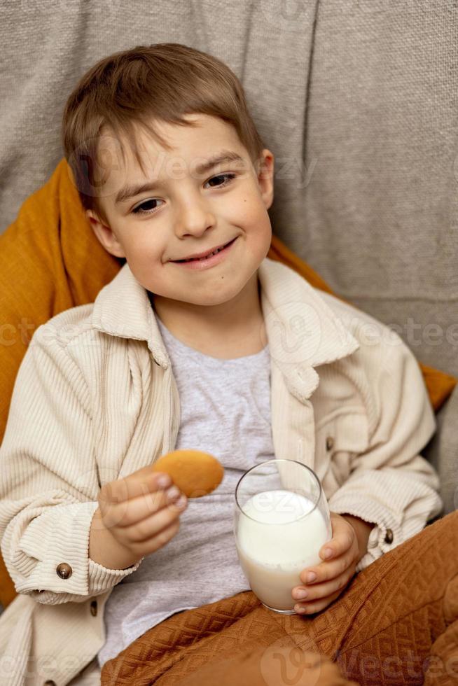 Little adorable boy sitting on the couch at home, drinking milk with cookie. Fresh milk in glass, dairy healthy drink. Healthcare, source of calcium, lactose. Preschool child with casual clothing. photo