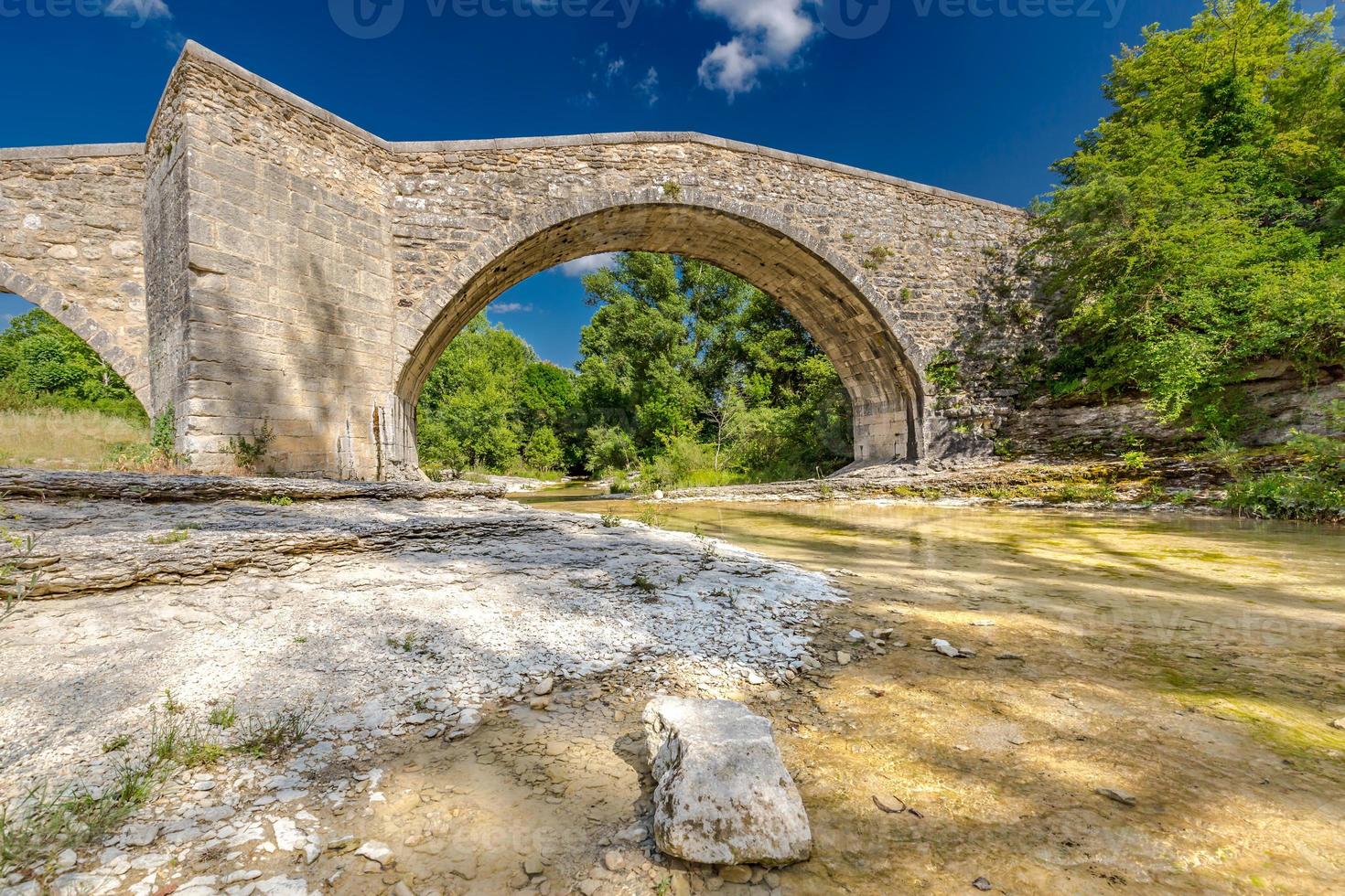 viejo puente de piedra bajo un cielo azul con nubes, pueblo histórico en francia, provence. arroyo tranquilo con rocas y árboles foto