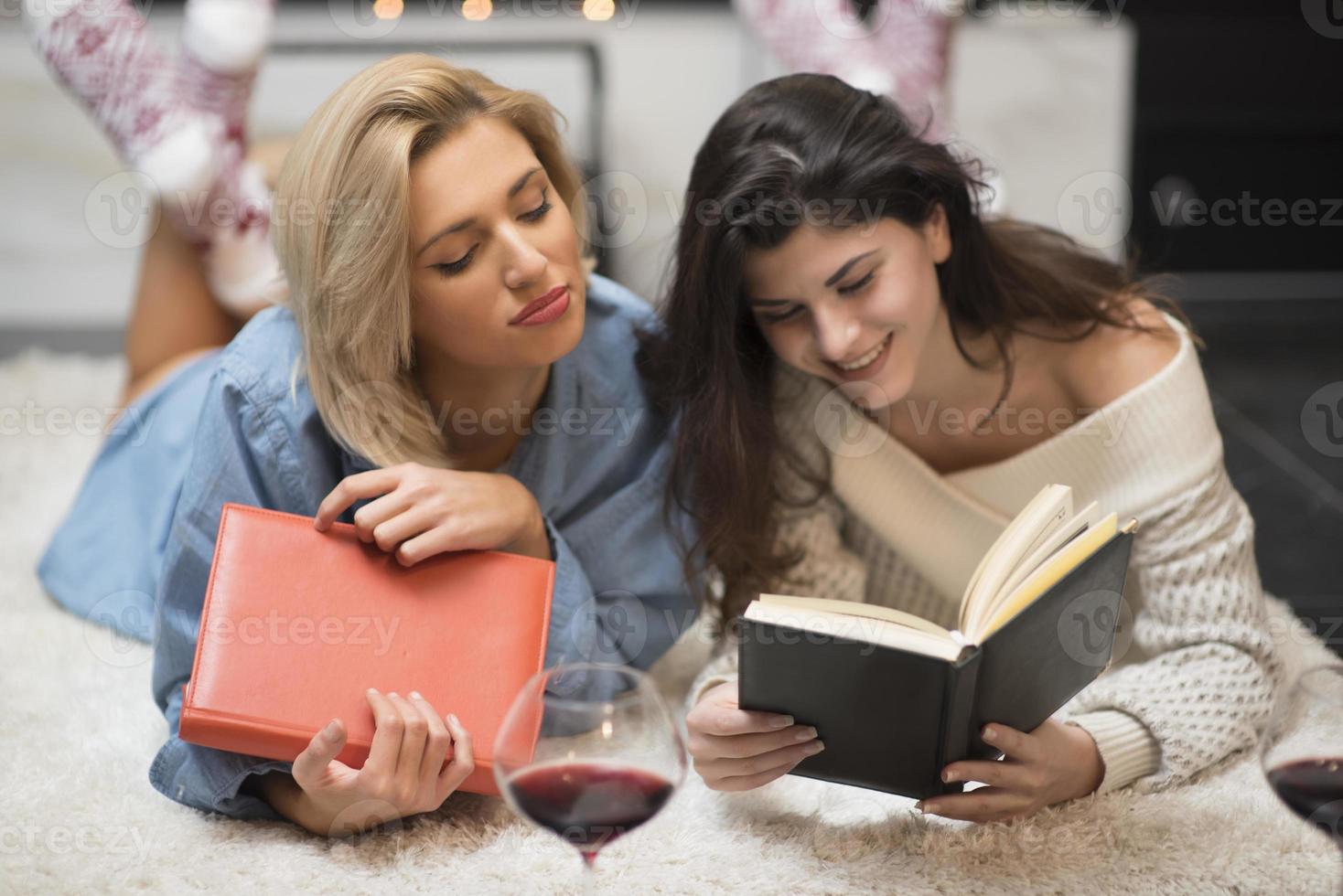 two female friends reading a book and drinking red wine by a fire place. life stile concept. photo