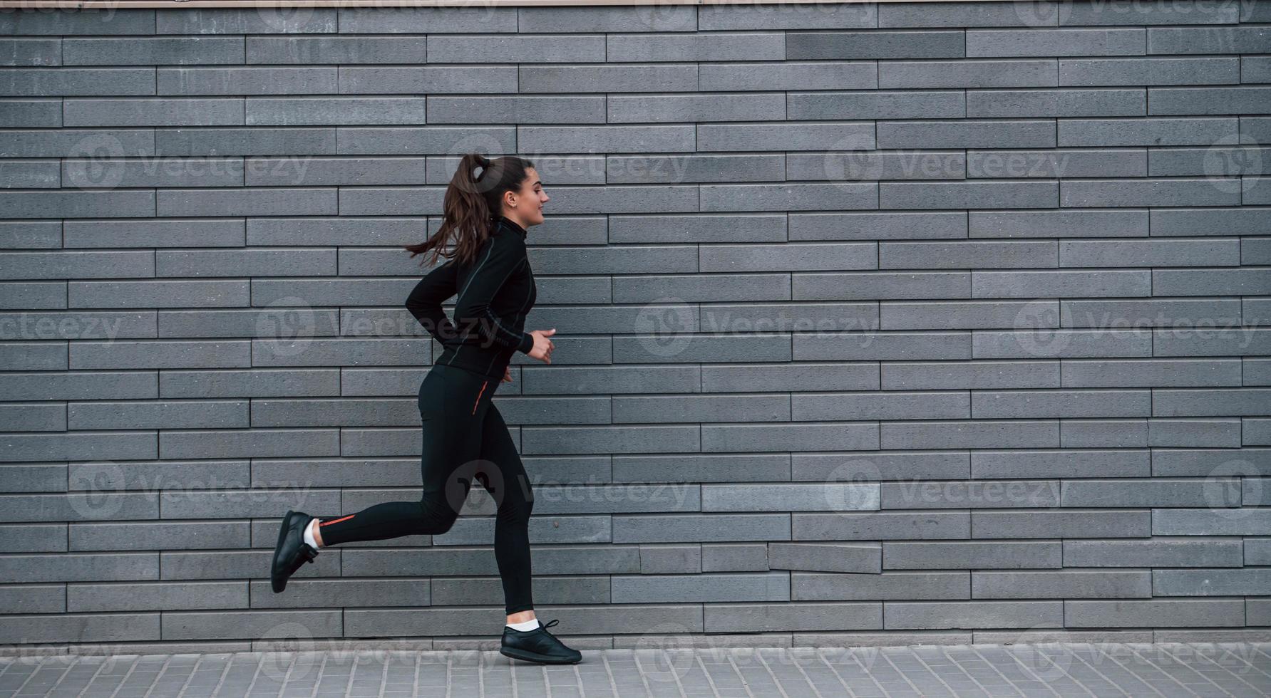 joven deportista con ropa deportiva negra corriendo al aire libre cerca de la pared gris foto