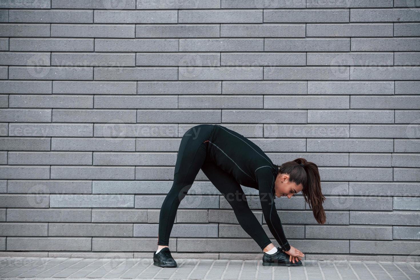 Young sportive girl in black sportswear outdoors doing stretching near gray wall photo